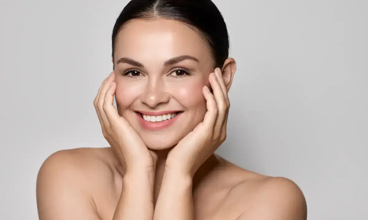 Uśmiechnięta kobieta z idealną, lśniącą cerą typu "glass skin", delikatnie dotykająca policzków dłońmi.